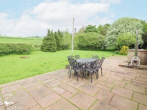 Eine Terrasse mit rundem Tisch und Stühlen, umgeben von einem mit Steinplatten belegten Bereich mit Blick auf ein grünes Feld und Bäume.
