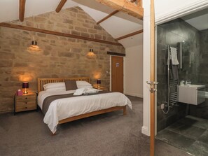 A bedroom with a stone wall, wooden beams, a bed with white bedding, bedside lamps, and a bathroom with a glass shower. - Miller's Wheel (Harrogate)