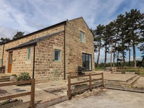 Un bâtiment en pierre avec une clôture en bois, une zone pavée agrémentée de parterres de fleurs et des arbres en arrière-plan.