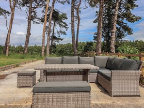 A wicker outdoor seating area with a sofa and ottomans, surrounded by tall trees and a wooden fence.