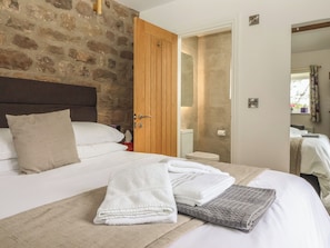Habitación de hotel con pared de piedra, puerta de madera, baño con inodoro y lavamanos, y cama con ropa blanca y toallas.