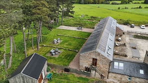 Un bâtiment en pierre doté d’un toit en pente, une cour clôturée avec des sièges extérieurs, et une vue sur un champ verdoyant.