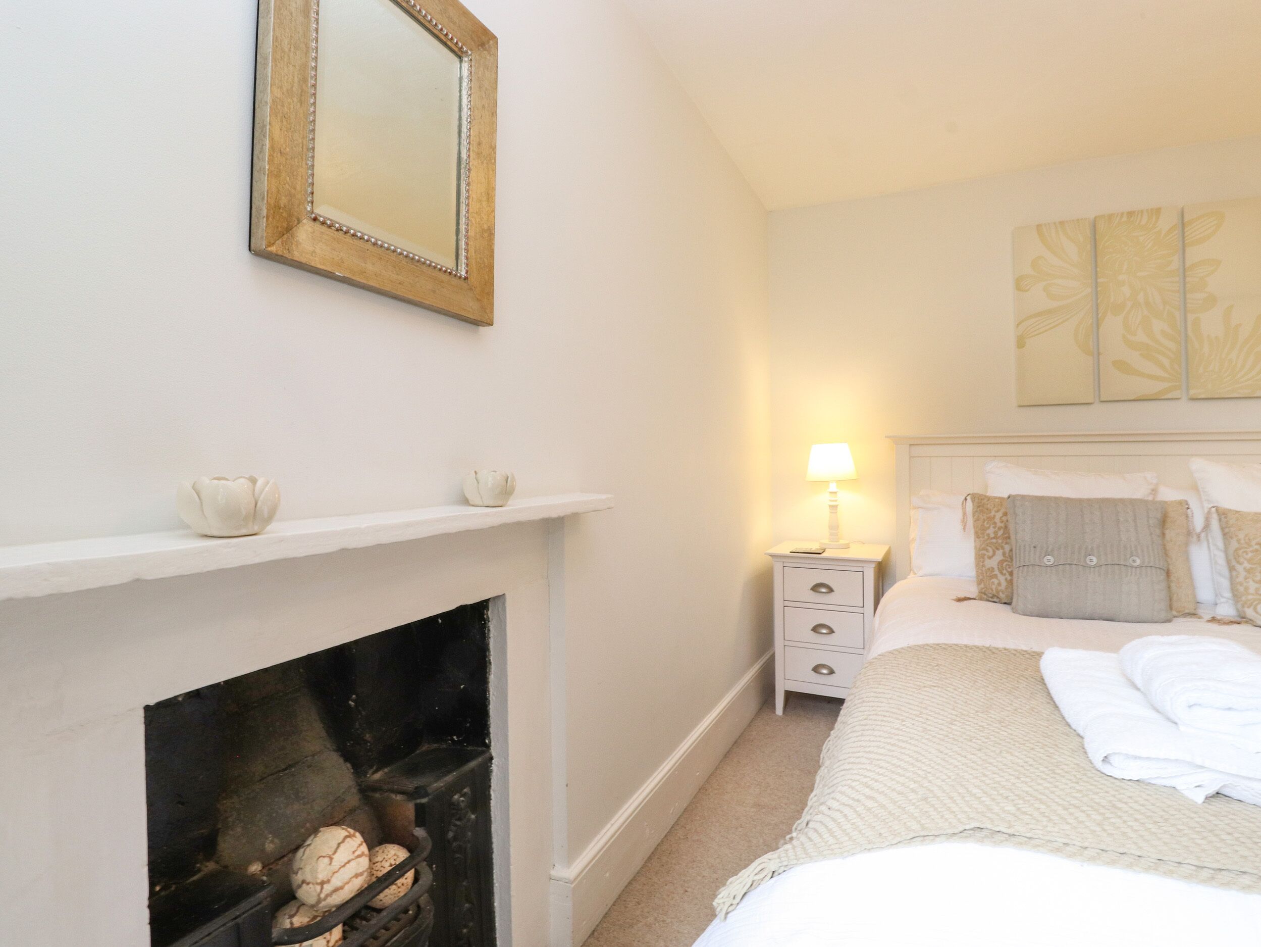 A bedroom with a fireplace, a bedside table with a lamp, a bed with pillows, and a wall-mounted mirror.