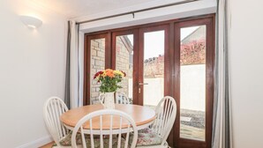Un comedor con una mesa de madera y cuatro sillas blancas, un jarrón con flores y vistas a un espacio exterior con pared de ladrillo y follaje rojo.