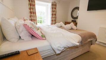 A neatly made bed with white linens, a remote control on a wooden side table, and a window with floral curtains.