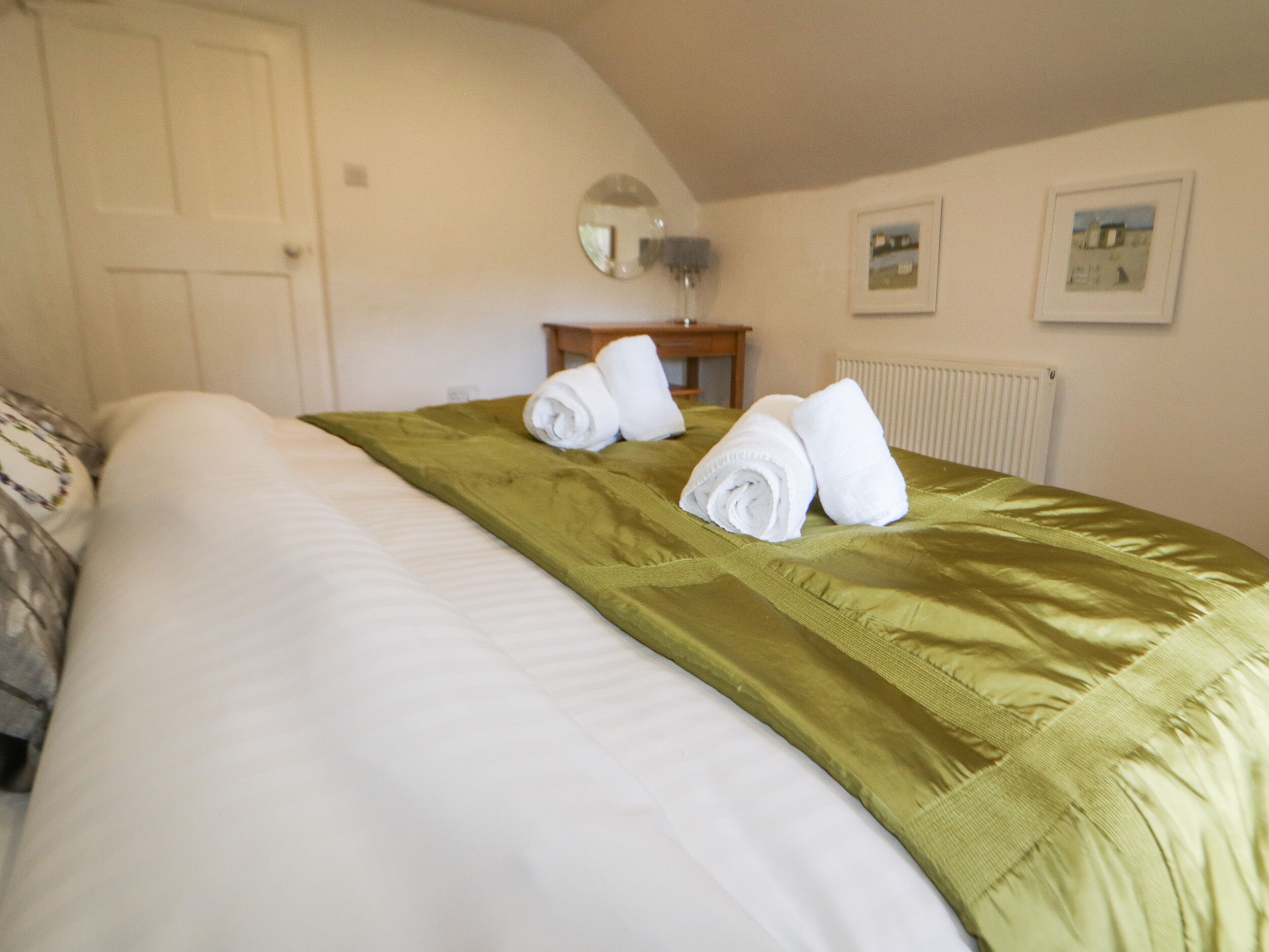 A neatly made bed with white linens and a green bedspread, a wooden nightstand with a lamp, and a framed picture on the wall.
