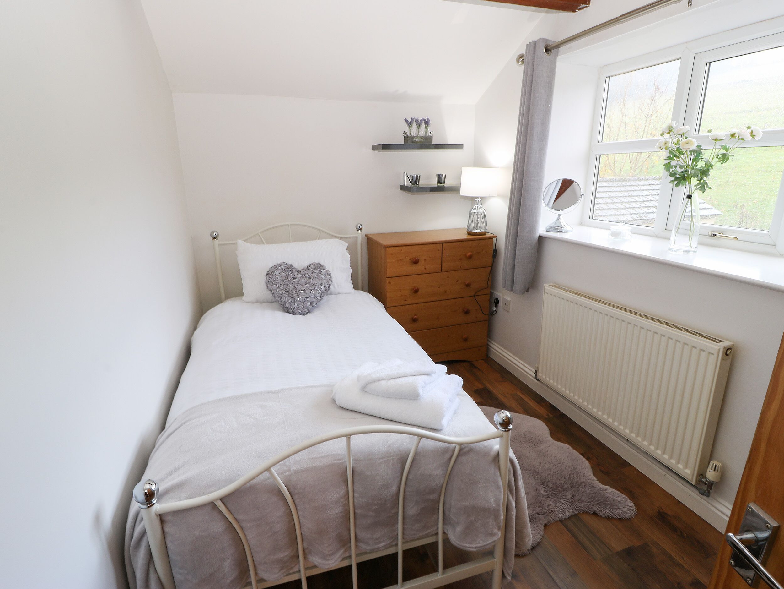 Un dormitorio ordenado con cama, cómoda, mesita de noche con lámpara, ventana con vistas y radiador.