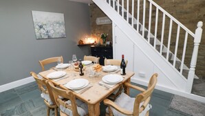 A dining area with a wooden table set for six, surrounded by six wooden chairs with cushioned seats. A staircase with a white railing is visible in the background. - The Coach House (Bradford)