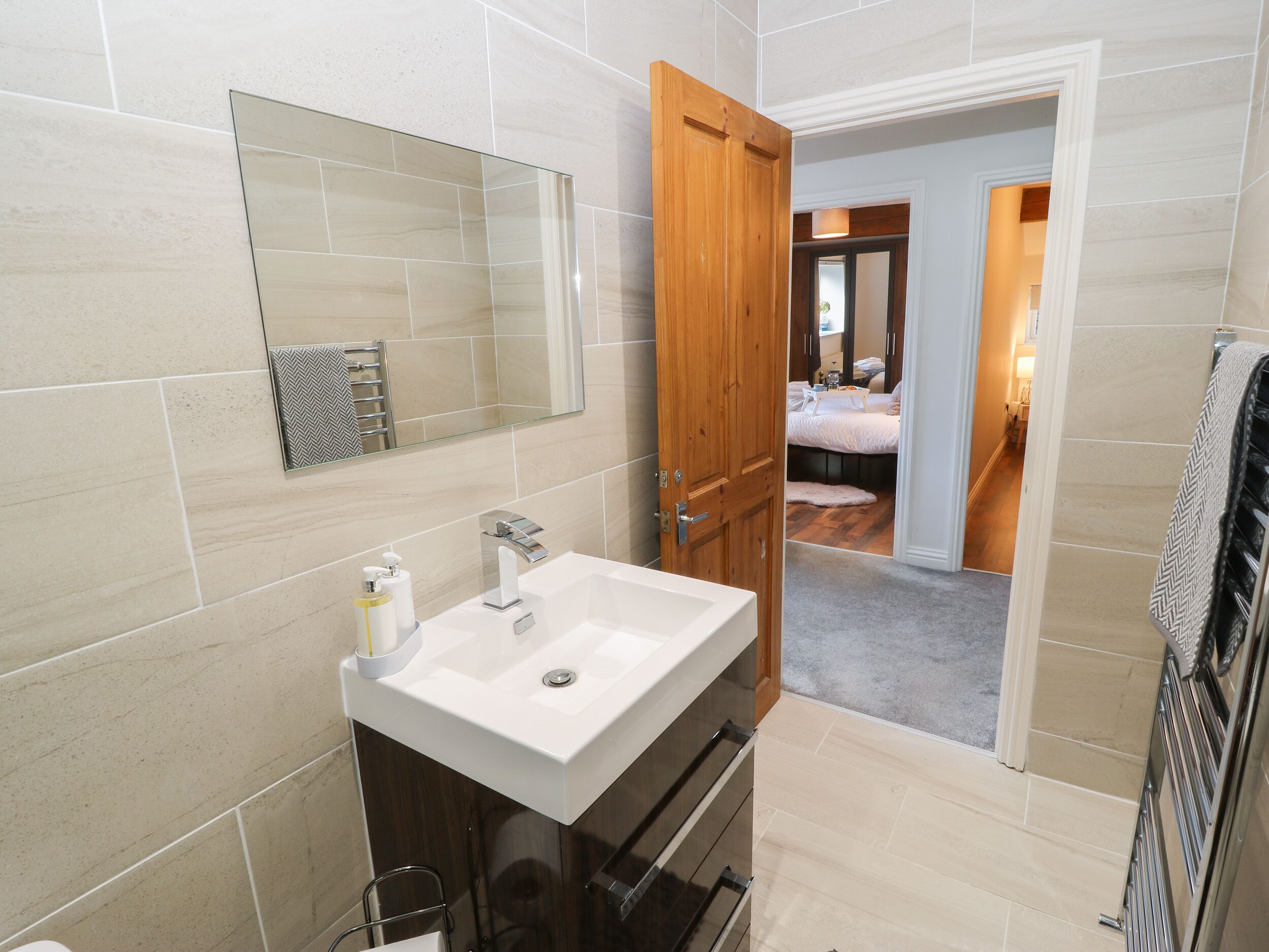 Un baño moderno con un espejo grande, un lavamanos blanco y una puerta de madera que da a un dormitorio.