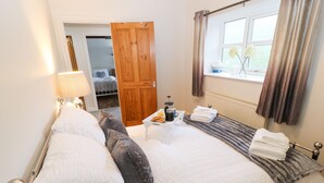 A neatly made bed with pillows, a bedside table with a cup and a book, a window with curtains, and a wooden door. - The Coach House (Bradford)
