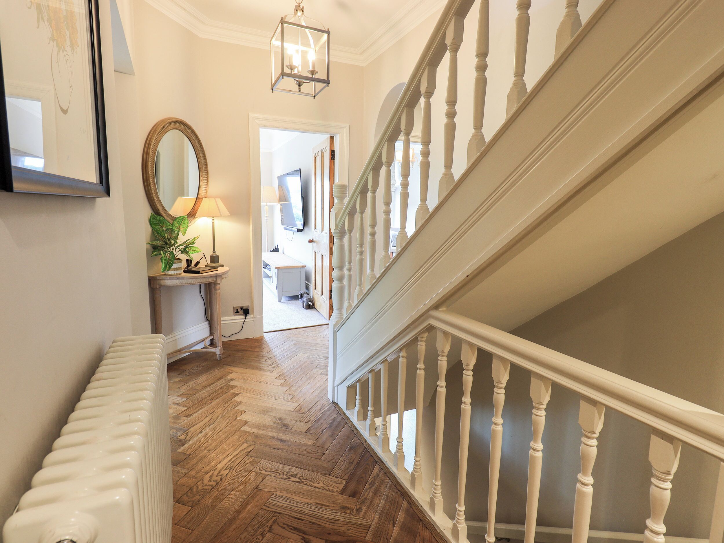 A hallway with a wooden floor, a radiator, a lamp, a mirror, and a staircase with white balusters.