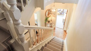 Eine Treppe mit weißen Geländern führt zu einer zweiten Etage. Darüber an der Wand hängt ein Spiegel, und im Hintergrund ist ein kleiner Tisch mit einer Lampe zu sehen.