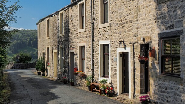 A stone building with multiple windows and doors, a paved road, and potted plants. - Winskill Cottage (Settle)