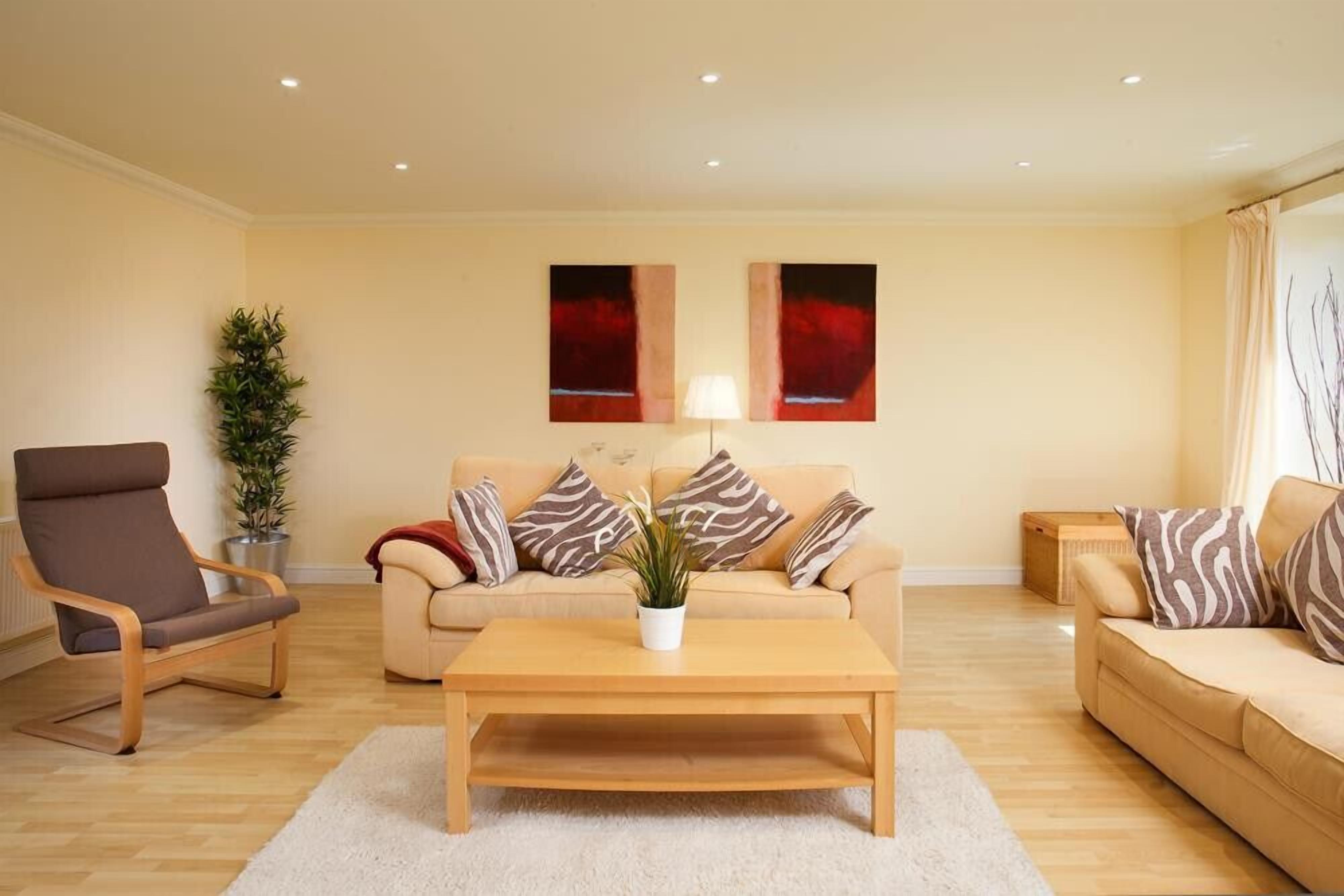 A modern living room with a beige sofa, a wooden coffee table, and a brown armchair. There are two abstract paintings on the wall and a potted plant on the floor.