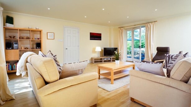 A spacious living room with two beige sofas, a wooden coffee table, and a TV stand. There is a bookshelf with various items and a door leading to an outdoor area. - The Old Barn (Windermere)