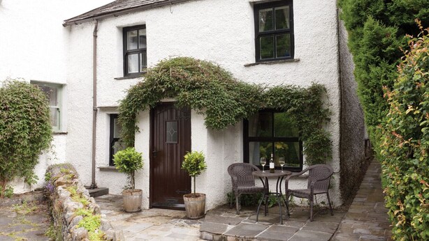 A white building with a brown door, two windows, and a small patio with chairs and a table. - Burrow Cottage (Windermere)