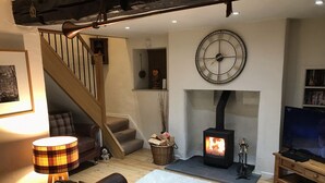 A cozy living room with a fireplace, a large clock, a staircase, and a television. - Burrow Cottage (Windermere)