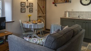 A cozy living room with a grey sofa, a small dining table with chairs, a television, and a kitchen area with a sink and cabinets. - Crooks (Axbridge)