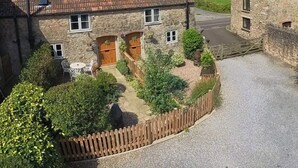 Une maison en pierre avec un toit recouvert de tuiles rouges, une porte en bois et un petit jardin entouré d’une clôture en bois.
