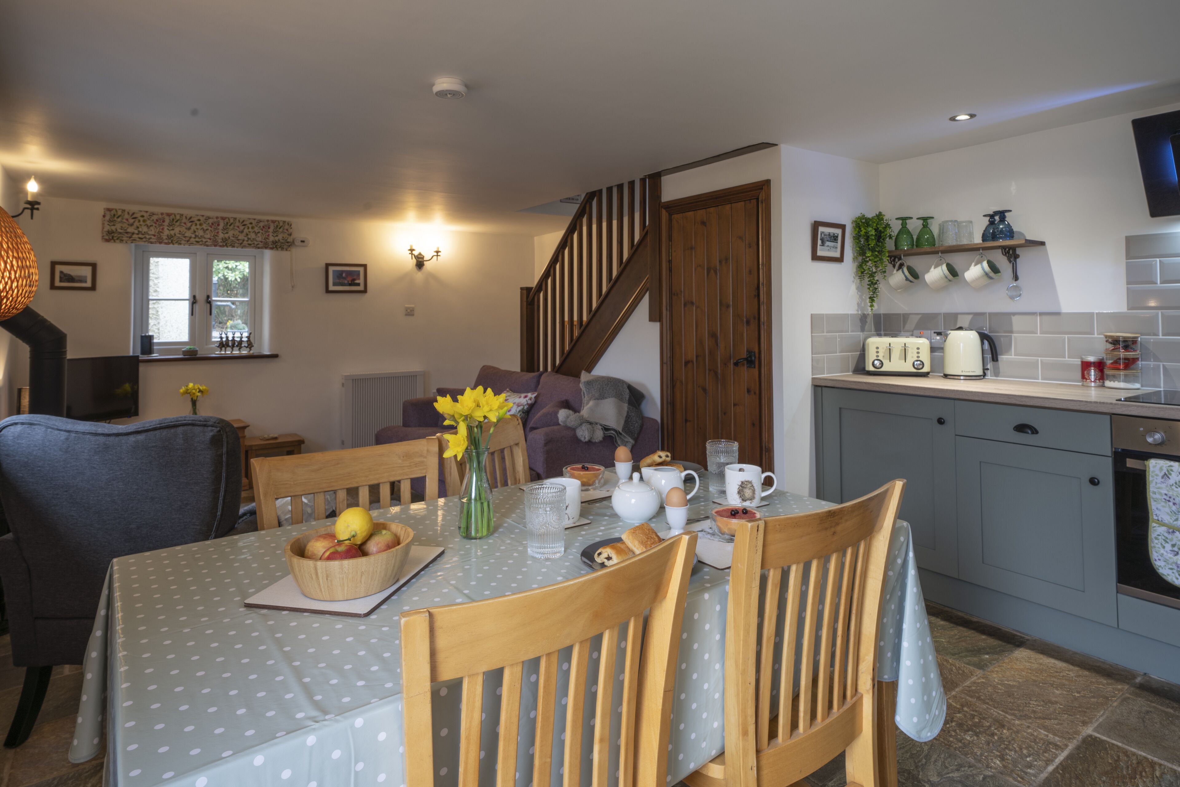 A dining area with a table set for four, a kitchen with grey cabinets, and a staircase leading to another level.