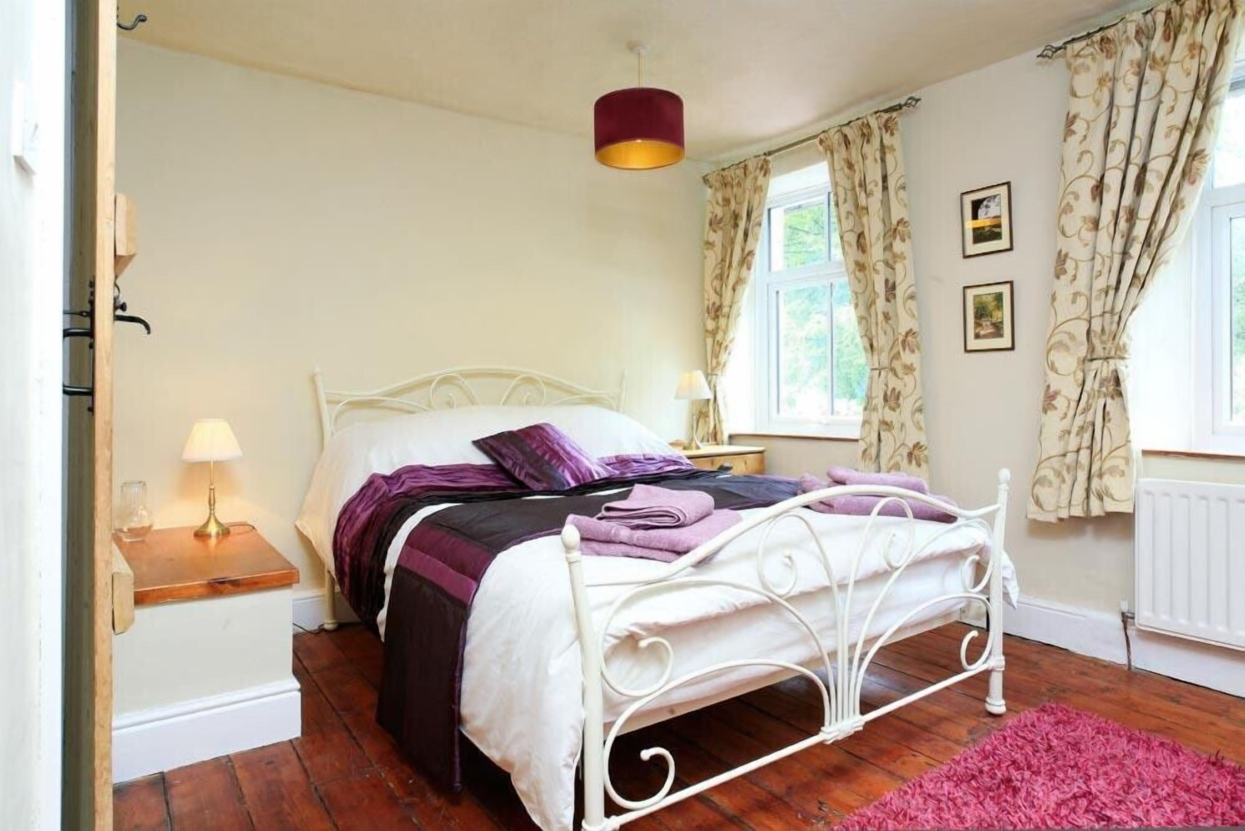 A bedroom with a white metal bed frame, a wooden nightstand with a lamp, a pink rug, and a window with floral curtains.