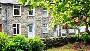 A stone building with multiple windows and a blue door, surrounded by lush greenery and pink flowers. - Stone Cottage at Staveley (Kendal)