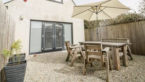 A patio with a wooden table and chairs, a beige umbrella, and a building with a sliding glass door. - Lily Cottage (Camelford)
