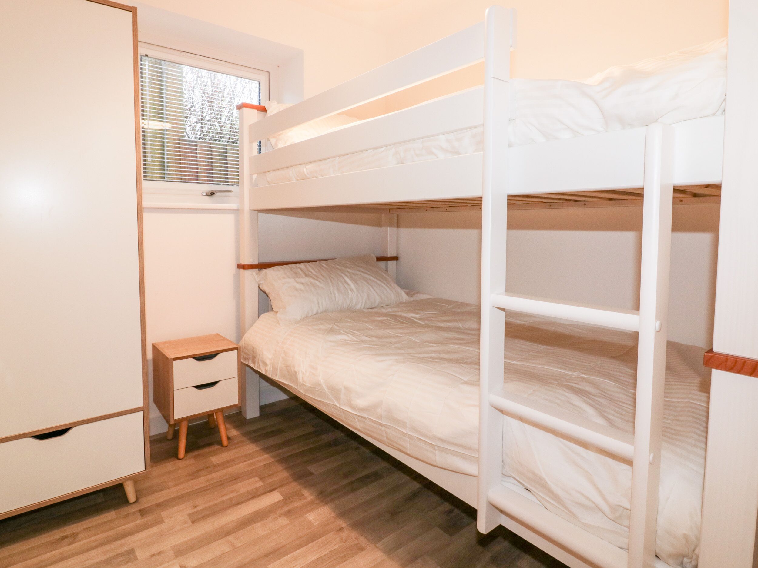 Quarto com beliche, uma pequena mesa de cabeceira de madeira, um guarda-roupa branco e uma janela com persianas.