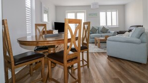 A modern living room with a dining area, featuring a round wooden table, wooden chairs, a light blue sofa, and a flat-screen TV.