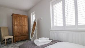 A bedroom with a bed, a wooden wardrobe, a chair, a mirror, and a window with blinds.