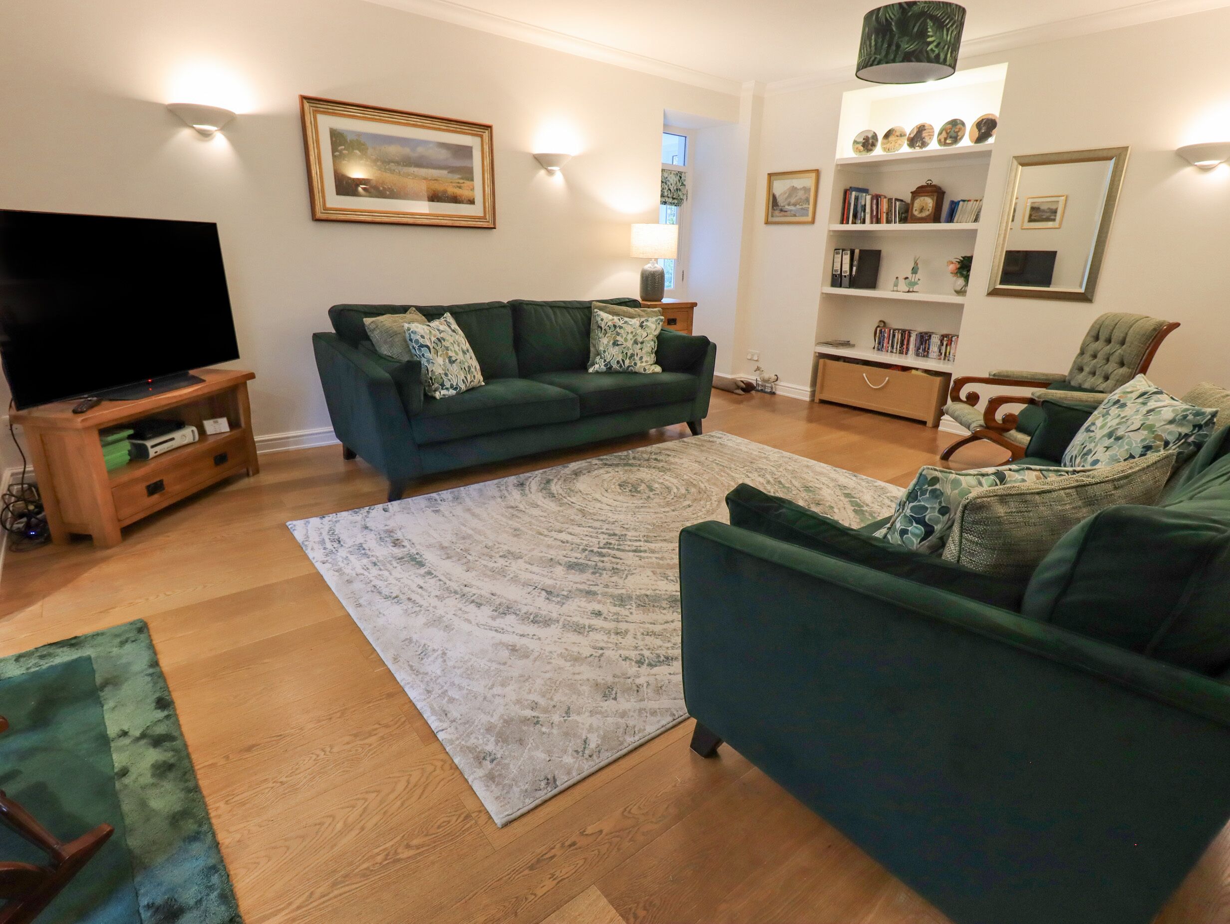 A living room with a green sofa, a flat-screen TV, a wooden entertainment unit, a patterned rug, and a bookshelf.
