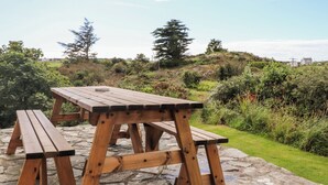 Una mesa de picnic de madera con bancos en un patio de piedra, rodeada de exuberante vegetación y un cielo despejado.