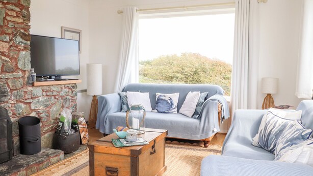 Una sala de estar acogedora con pared de piedra, un ventanal con cortinas, un sofá azul, una mesa de centro de madera y un televisor sobre una estantería.