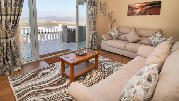 A living room with a sofa, a coffee table, and a view of the outdoors through a sliding glass door. - Mcauliffe Lodge (Borth)