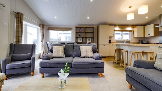 Een moderne woonkamer met een grijze zithoek, een bijzettafeltje met een vaas, en een keukenhoek met houten krukken.
