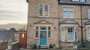 A two-story stone house with a blue front door, multiple windows, and a small porch area with potted plants. - Arlington House (Matlock)
