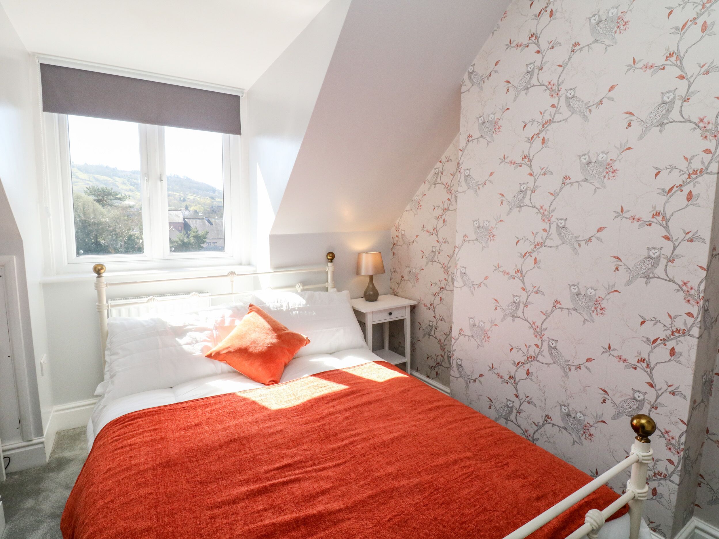 A bedroom with a bed, a nightstand, a lamp, a window, and a wall with a floral pattern.