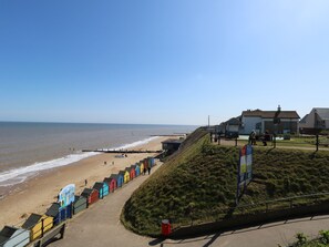 A coastal road with beach huts, a grassy hill, and a clear view of the sea. - The Nook (Norwich)