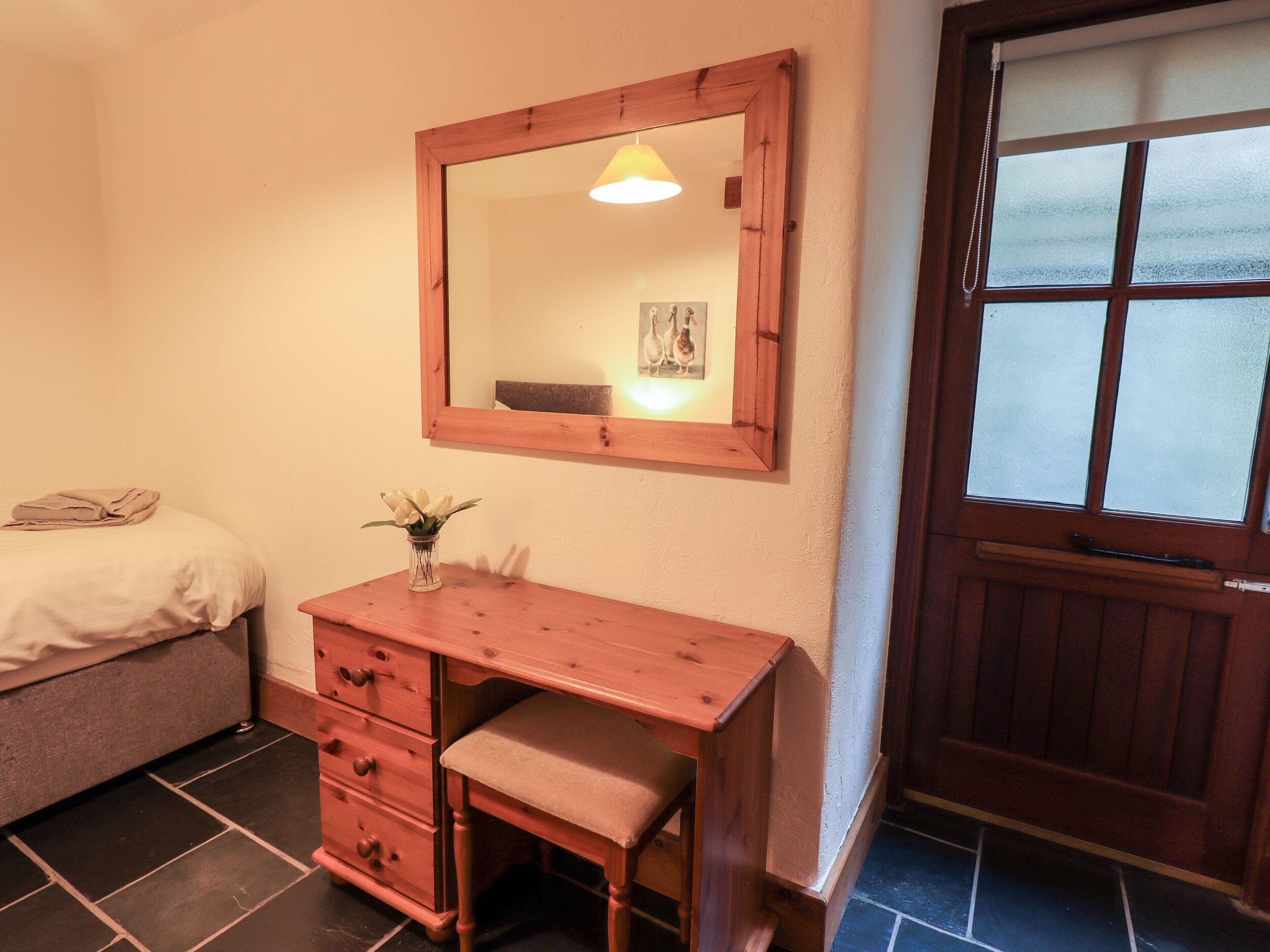 Une chambre avec une commode en bois, une table de chevet ornée d’un vase de fleurs, un lit avec une serviette pliée, un miroir avec un cadre photo et une fenêtre à vitres dépolies.