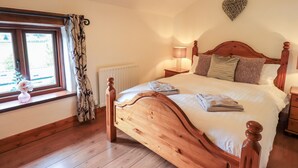 A bedroom with a wooden bed, a bedside table with a lamp, a window with curtains, and a radiator. - Millrace Cottage (Kendal)