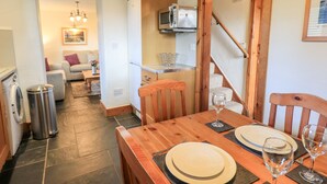 A dining area with a wooden table set for two, a microwave above the counter, and a staircase leading to another level. - Millrace Cottage (Kendal)