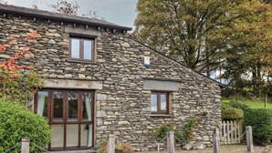 A stone house with two windows, a wooden fence, and a paved driveway. - Millrace Cottage (Kendal)