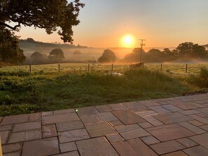 A paved outdoor area with a view of a sunrise over a misty field. - The Cosy Cowshed (Axminster)