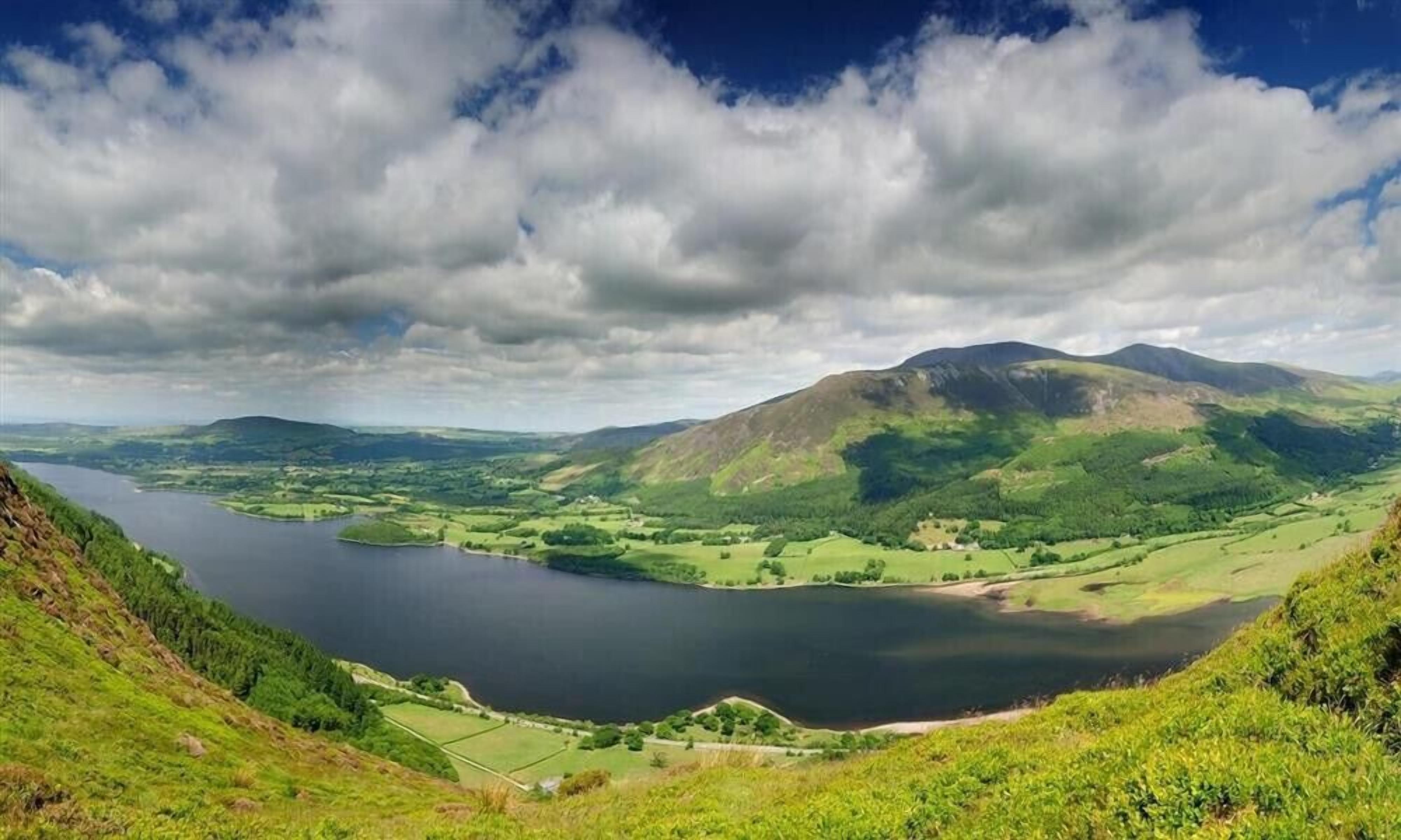 A panoramic view of a lake surrounded by green hills and mountains under a partly cloudy sky.
