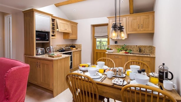 A kitchen with wooden cabinets, a dining table set for breakfast, and a red chair. - Barn Cottage (Cockermouth)