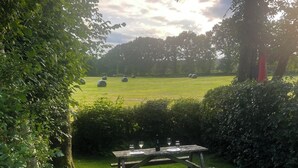 A picnic table with a bottle and glasses, surrounded by greenery and hay bales in the background. - Barn Cottage (Cockermouth)