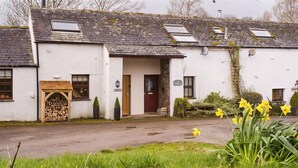 A white building with a traditional design, featuring a wooden door, a small porch with a bench, and a stack of firewood. - Barn Cottage (Cockermouth)