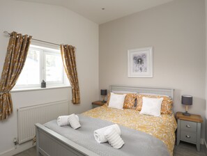 Une chambre bien rangée, avec un lit, deux tables de chevet, une fenêtre avec des rideaux, un radiateur et un tableau encadré au mur.