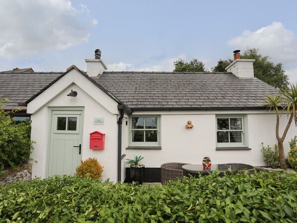 Une petite maison blanche avec une porte verte, une boîte aux lettres rouge et un petit jardin agrémenté d’une table en osier et d’un objet décoratif.