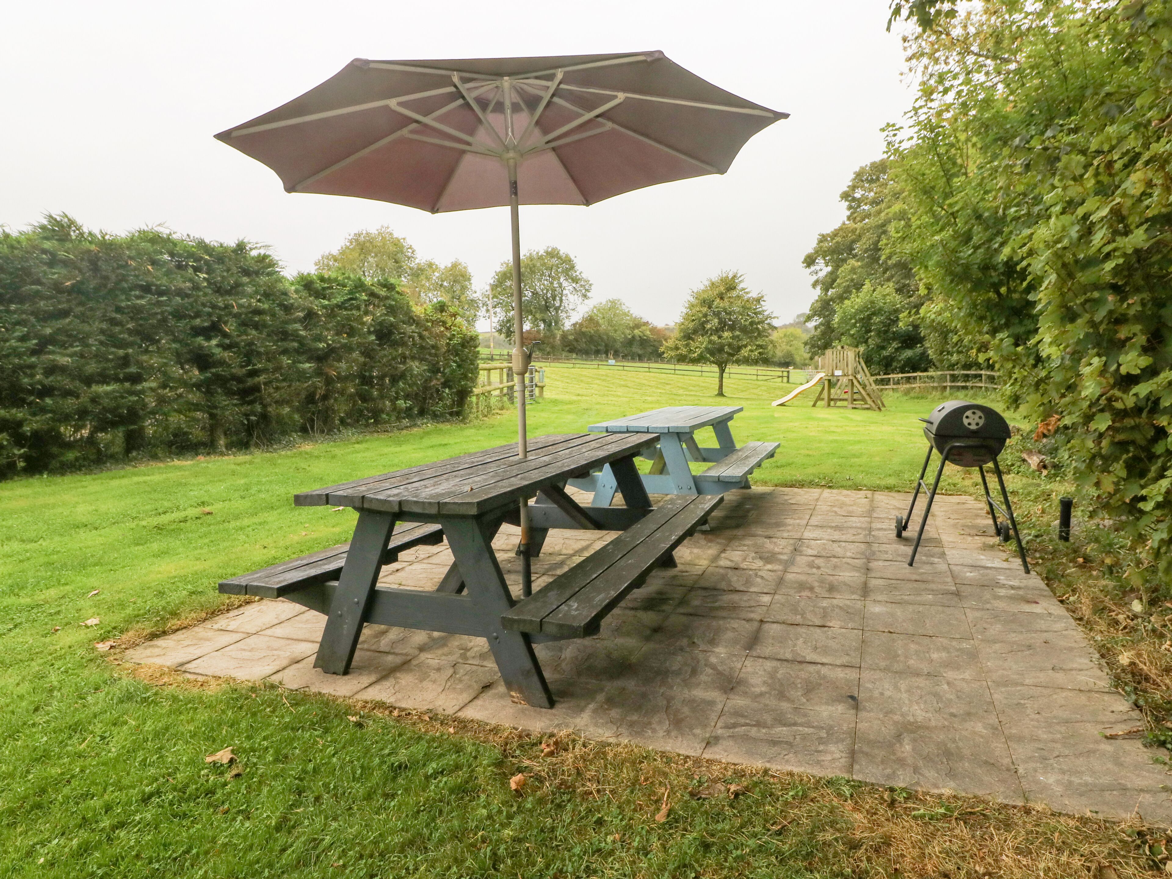 A picnic table with an umbrella, a barbecue grill, and a playground in the background.